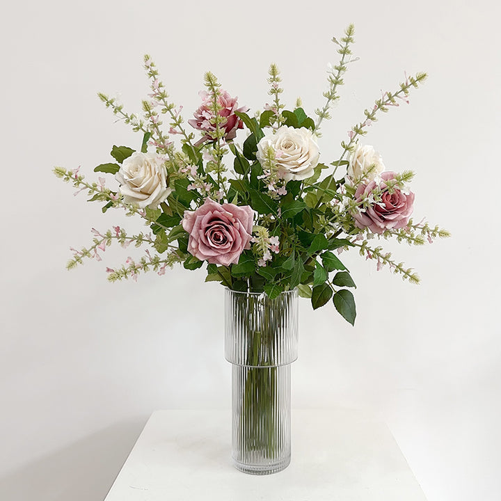 Dusty pink and cream faux rose bouquet with pale pink filler sprigs in a tall clear ribbed vase, isolated on a plinth.