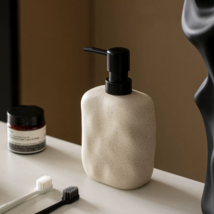 Cream speckled ceramic soap dispenser with a left-pointing black pump standing on a vanity with toothbrushes.