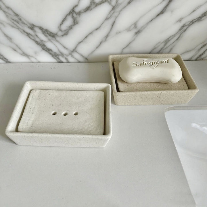 Two cream speckled ceramic soap dishes on a counter, one empty showing three drain holes and one holding a bar of soap.