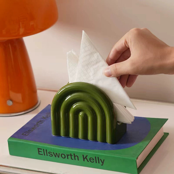 Hand placing folded white napkins into a glossy olive green rainbow ceramic holder on stacked books