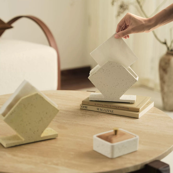 Hand placing a folded napkin into a white speckled geometric ceramic napkin holder on a wood table.