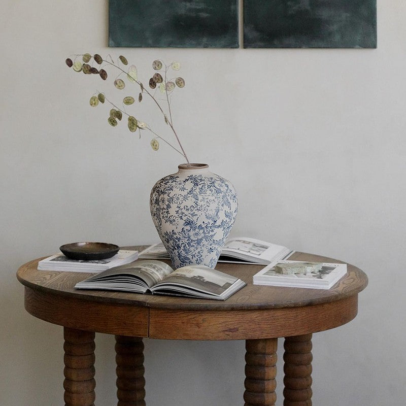 Blue and white porcelain vase with dried branches on round wooden table with open books and bowl