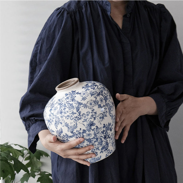 Person wearing dark blue dress holding large blue and white floral ceramic vase indoors near green plant