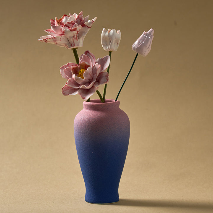 Porcelain peony diffuser set with two open pink blooms and two buds in a pink-to-blue ombre vase on a tan background.