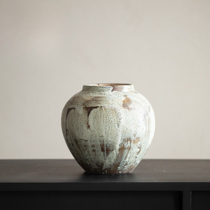 Round squat ceramic vase with mottled cream-green speckled glaze and narrow neck on a black console.
