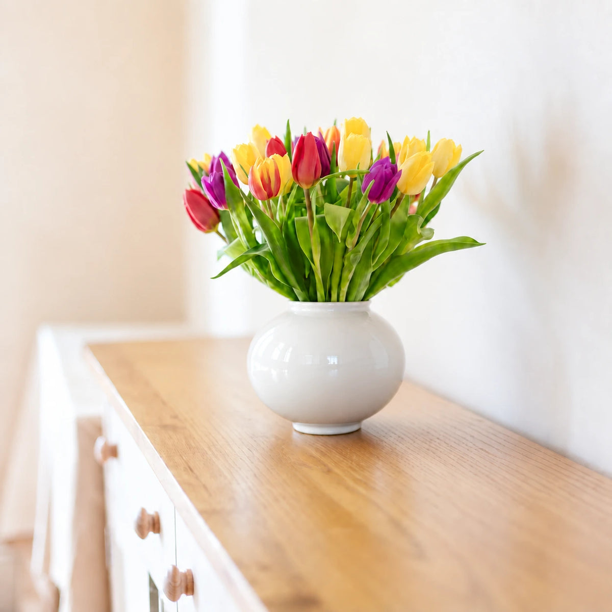 Small glossy white moon vase holding colorful tulips on a white sideboard.