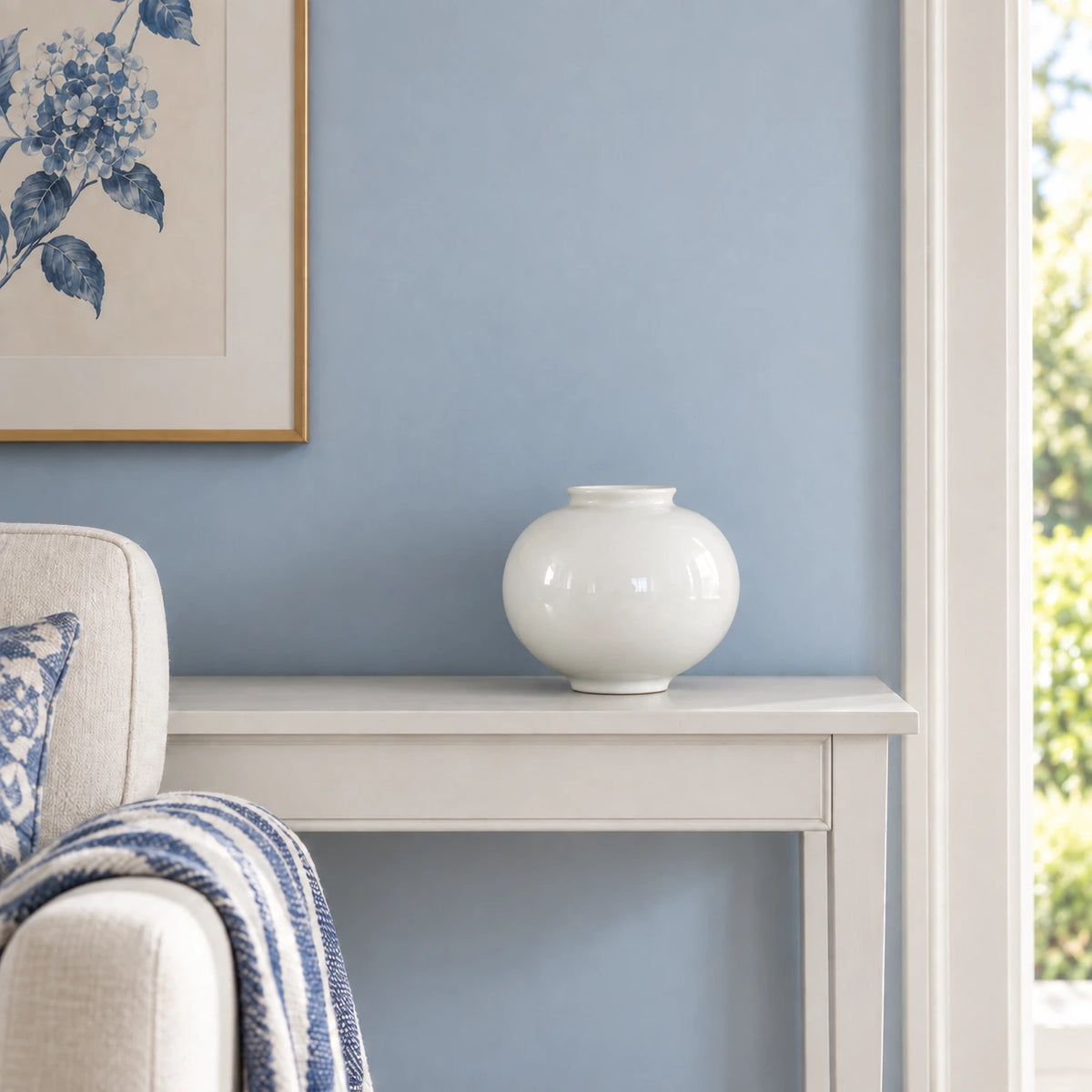 Empty small white moon vase on a painted console beside blue and white upholstery.