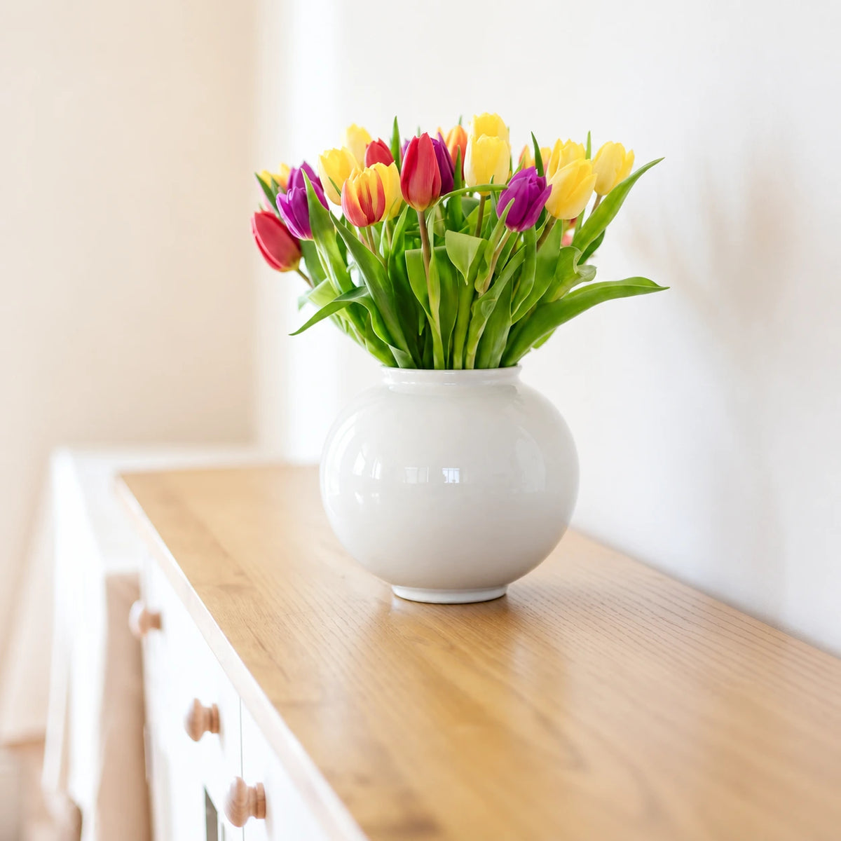 Medium glossy white moon vase holding colorful tulips on a white sideboard.