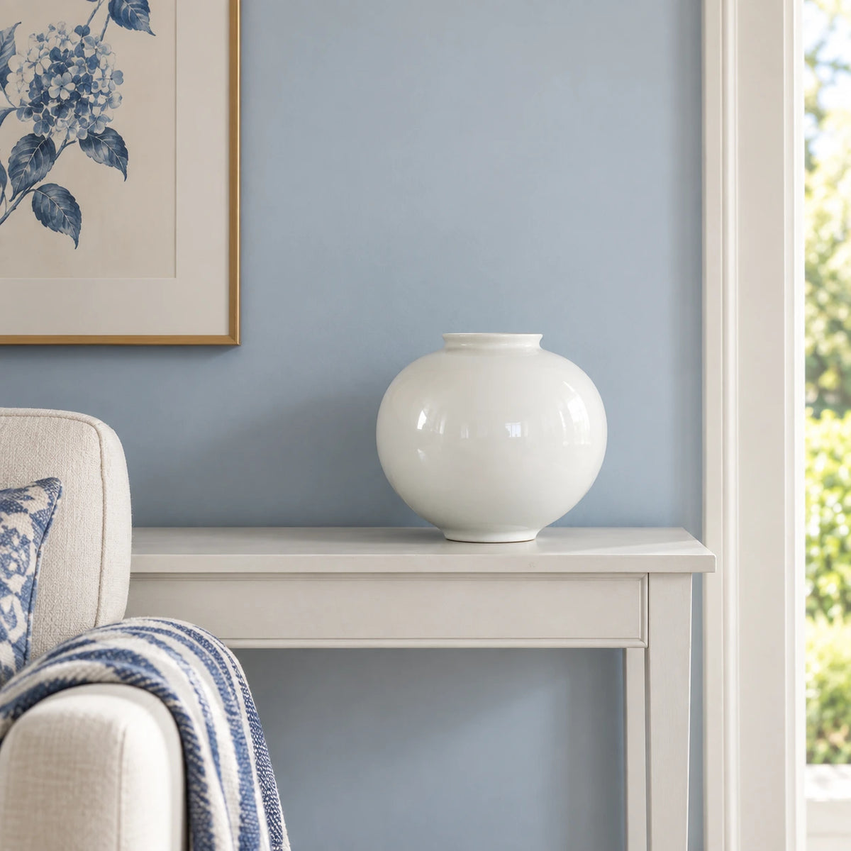 Empty medium white moon vase on a painted console beside blue and white upholstery.