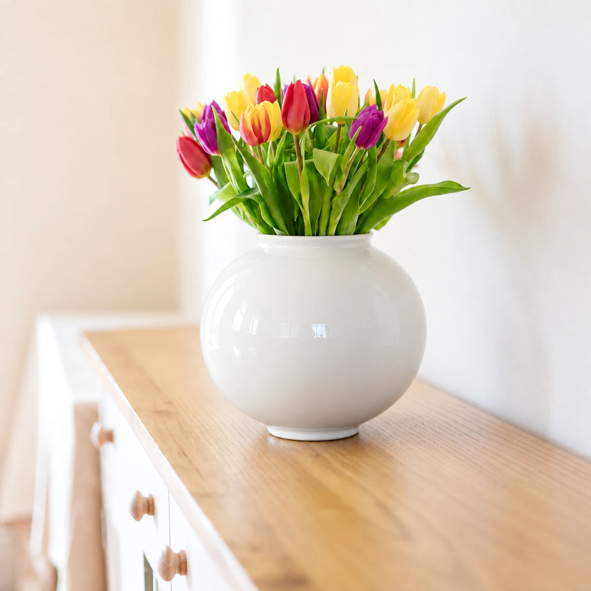 Large glossy white moon vase holding colorful tulips on a white sideboard.