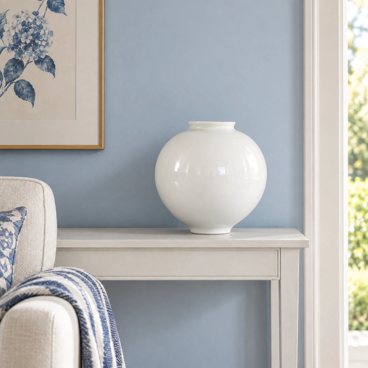 Empty large white moon vase on a painted console beside blue and white upholstery.