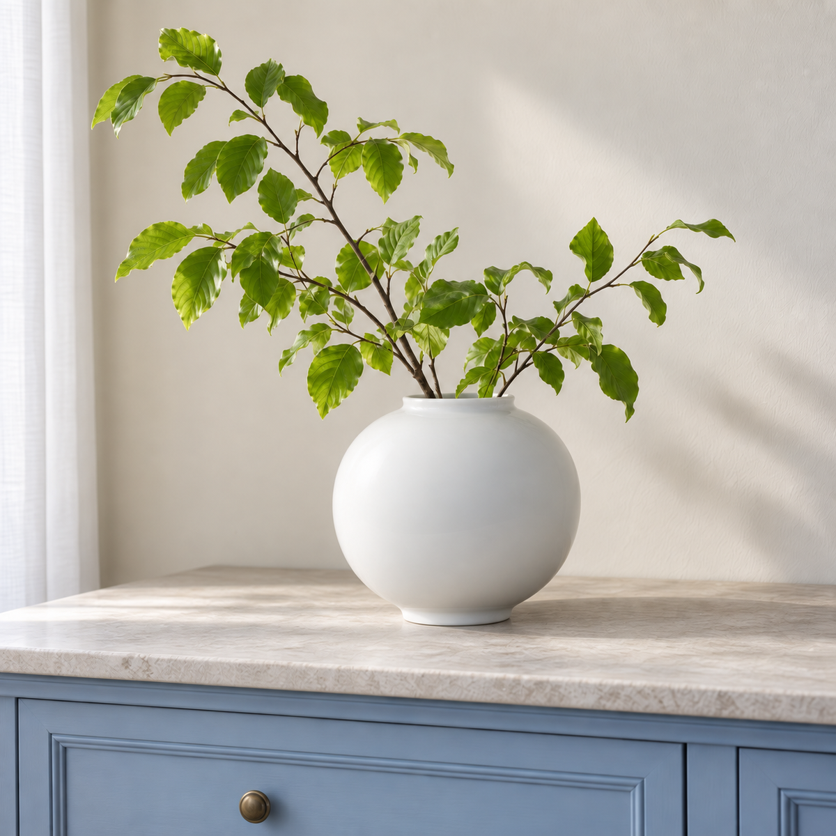 Medium white moon vase with leafy branches on a pale stone-topped blue sideboard.