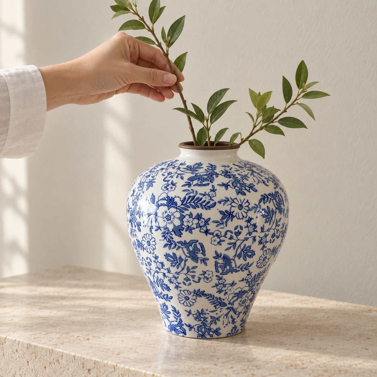 A hand arranging a leafy stem in a blue and white ceramic vase on a light stone surface.
