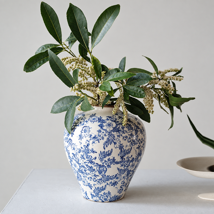 Blue and white floral ceramic vase with green leafy branches and small cream flowers on a pale tabletop in natural light.