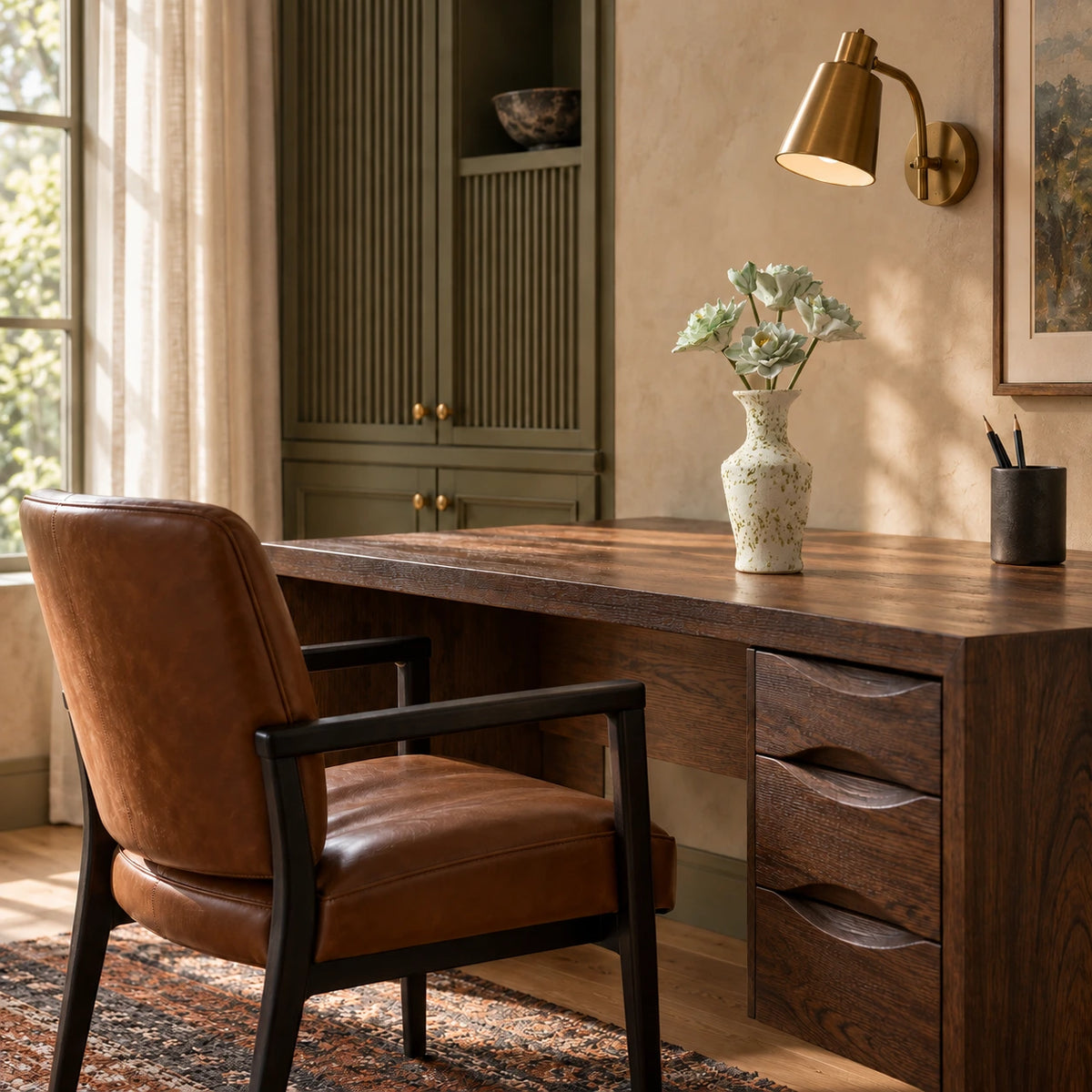 Five green-and-white ceramic flowers in a speckled cream vase on a dark oak desk with sculpted drawers, a leather chair and a brass wall reading light.