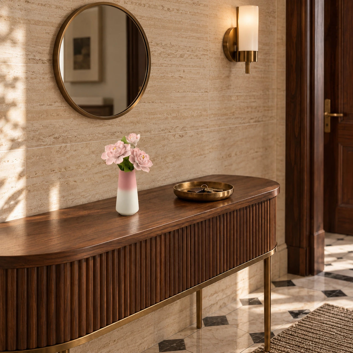 Three pink porcelain flowers in a pink-to-white tapered vase on a fluted walnut entry console against a travertine wall with a bronze-rim mirror.