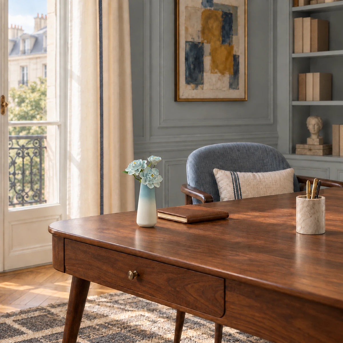 Three sky-blue porcelain flowers in a blue-to-white tapered vase on a walnut writing desk beside a blue-grey chair in a panelled apartment study.