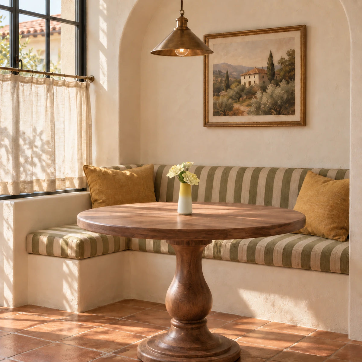 Three pale yellow porcelain flowers in a yellow-to-white tapered vase on a round wooden breakfast table with an olive striped banquette and terracotta floor.
