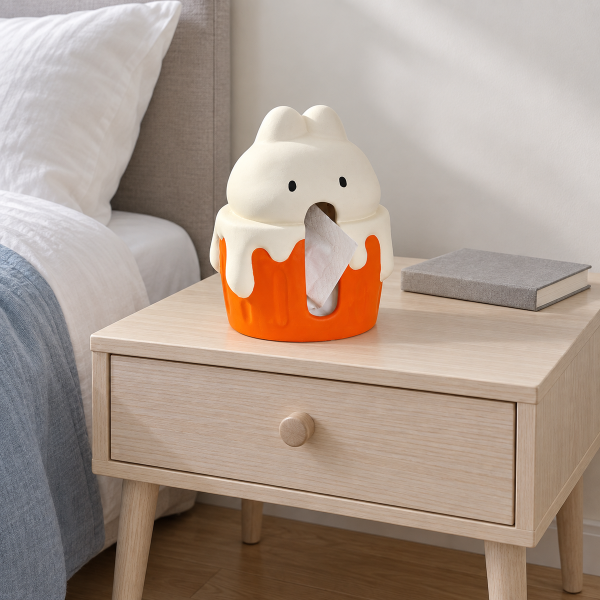 Cache-boîte à mouchoirs lapin cupcake sur une table de chevet en bois près de linge de lit blanc et d’un livre fermé
