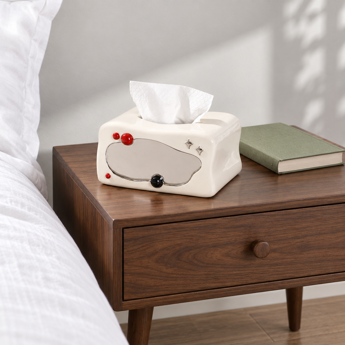 Ivory retro-TV tissue box cover on a wooden nightstand beside white bedding and a closed green book