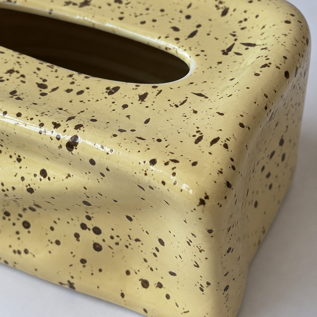 Close view of dark flecks and glossy wavy folds on the pale yellow ceramic tissue box cover.