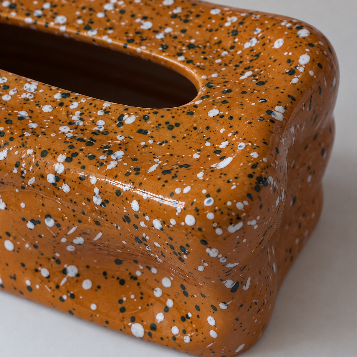 Close view of white and dark flecks across the glossy wavy folds of a terracotta tissue box cover.