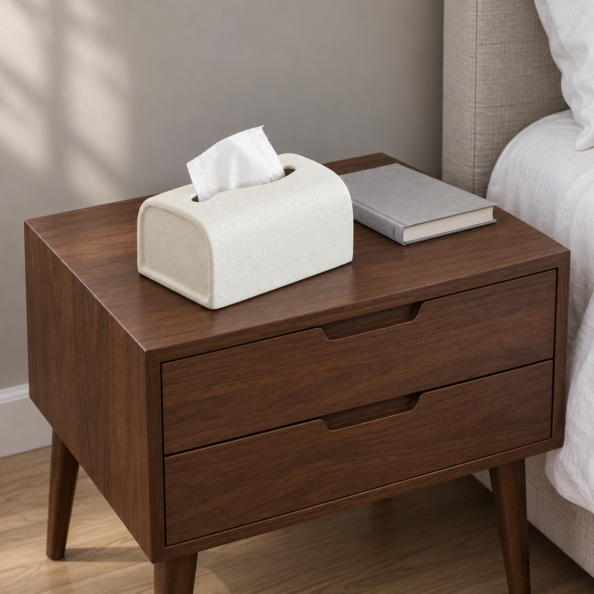 Textured ceramic tissue box cover on a walnut bedside table beside white bedding.