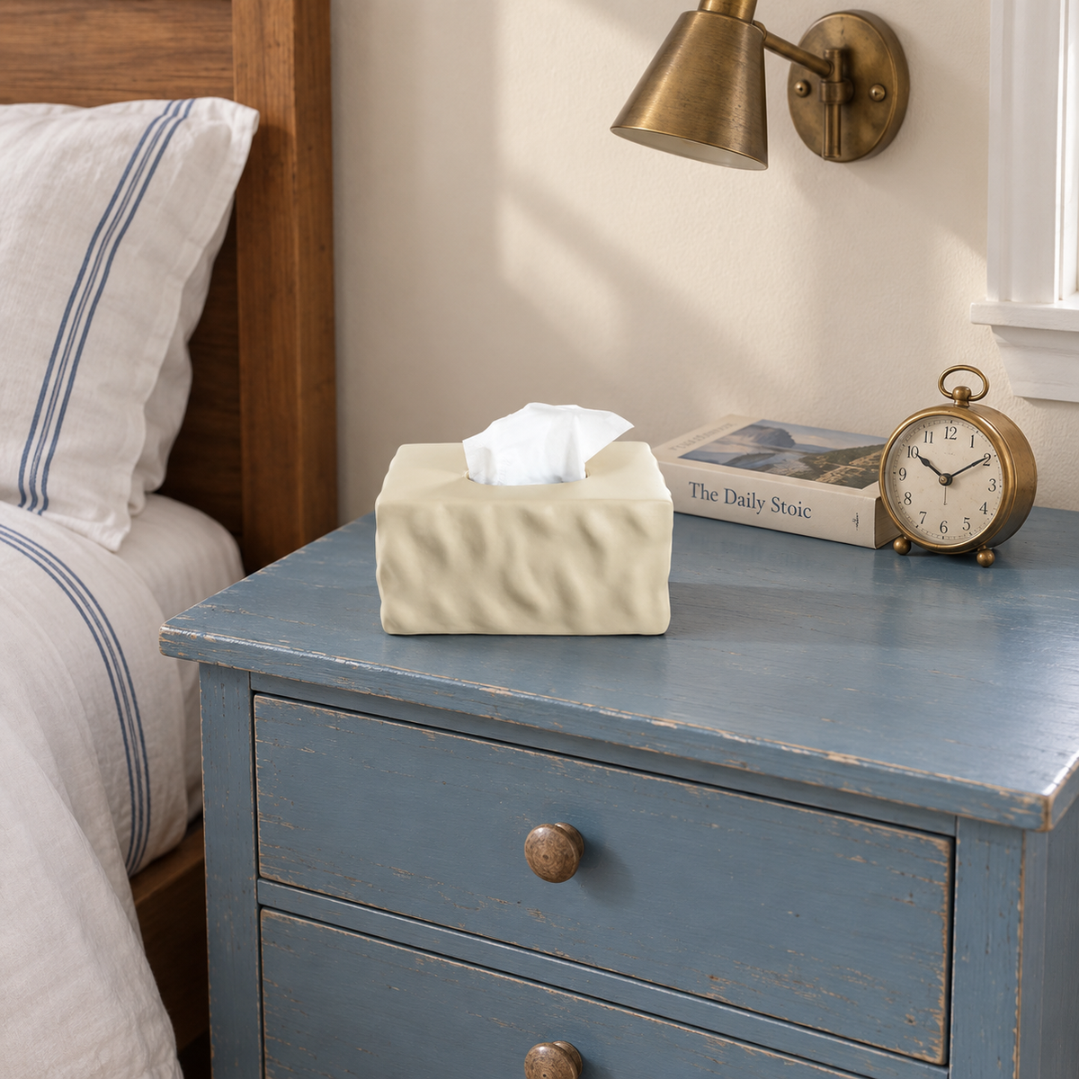 Light beige ceramic tissue box cover on a worn blue bedside chest with a book and small clock.