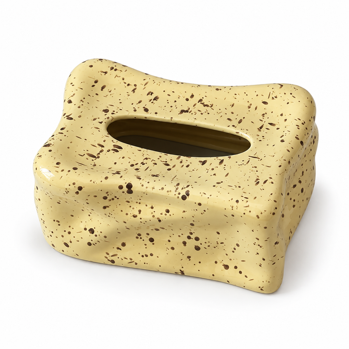 Pale yellow ceramic tissue box cover with dark speckles, two wavy folds and an empty oval top opening.