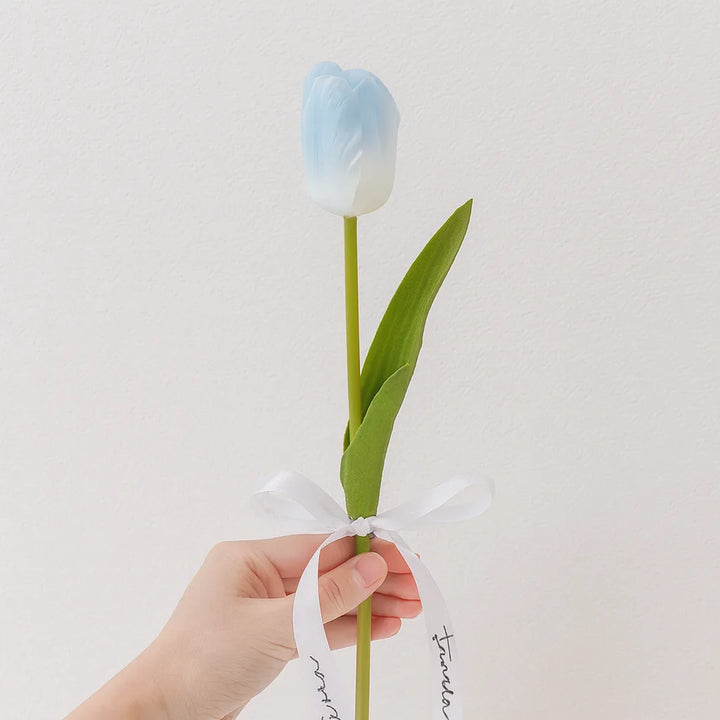 Single blue and white ombre faux tulip stem with two pointed leaves and a white tied ribbon, held by a hand.