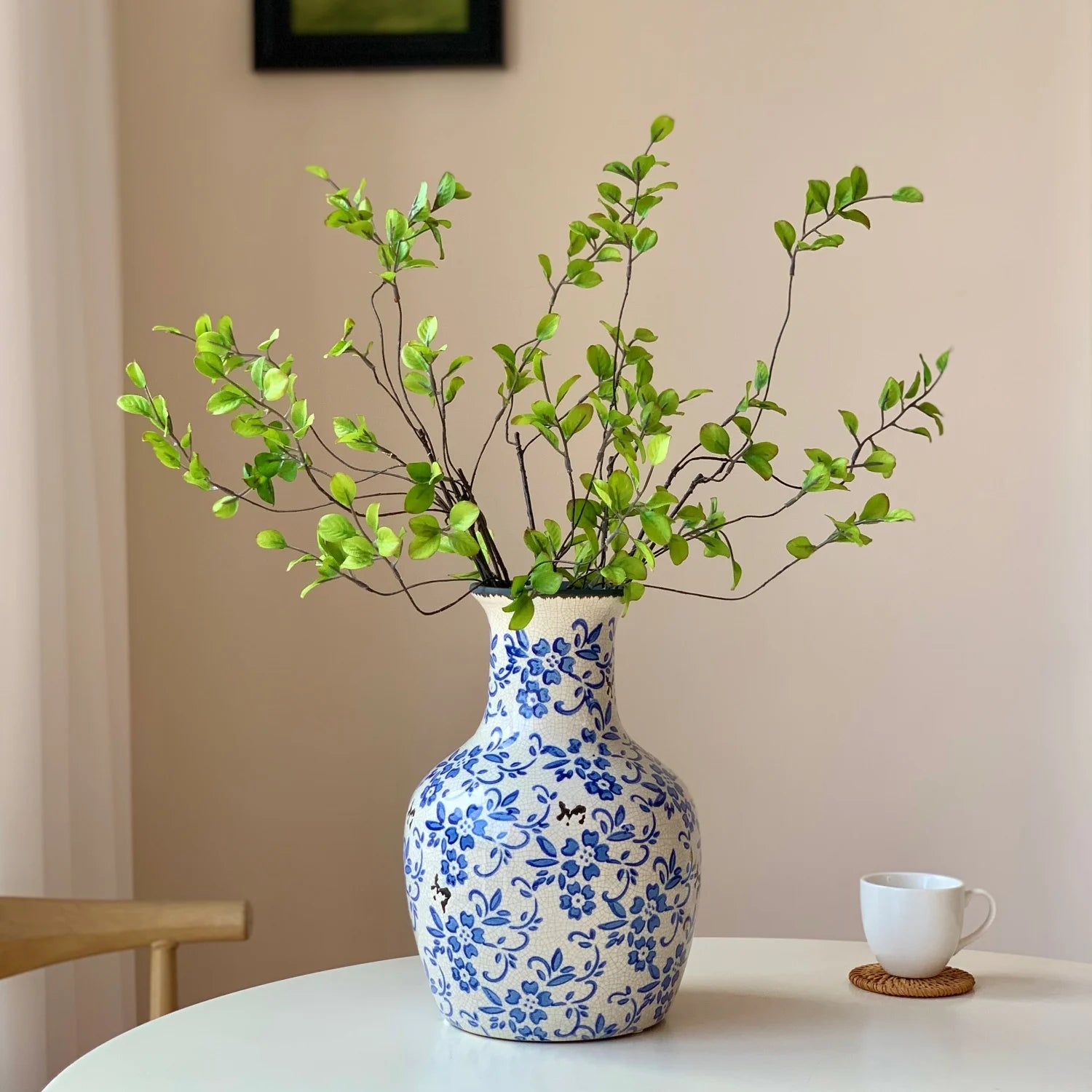 Decorative blue and white ceramic vase with green leafy branches on a white table with a white coffee cup