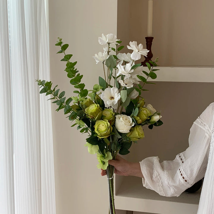 Hand holding a green-and-white faux bouquet of lime roses, white blooms, and eucalyptus foliage by a sheer curtain.