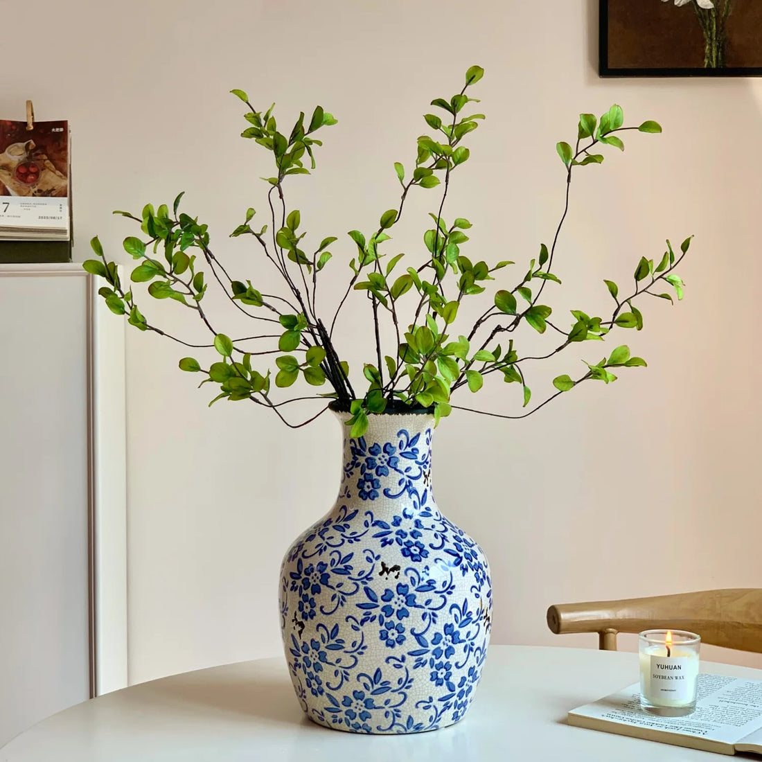 Blue and white floral ceramic vase with green leafy branches on white table next to lit soy candle