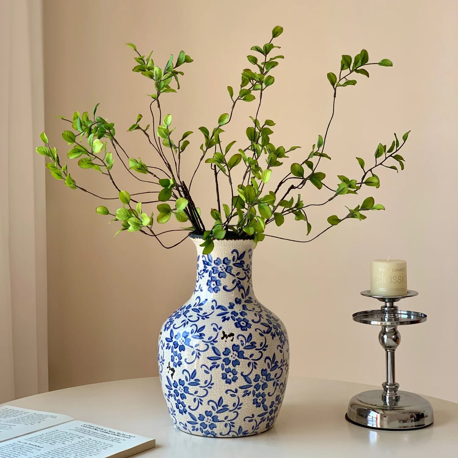 Blue and white floral ceramic vase with green branches on table next to silver candle holder with white candle