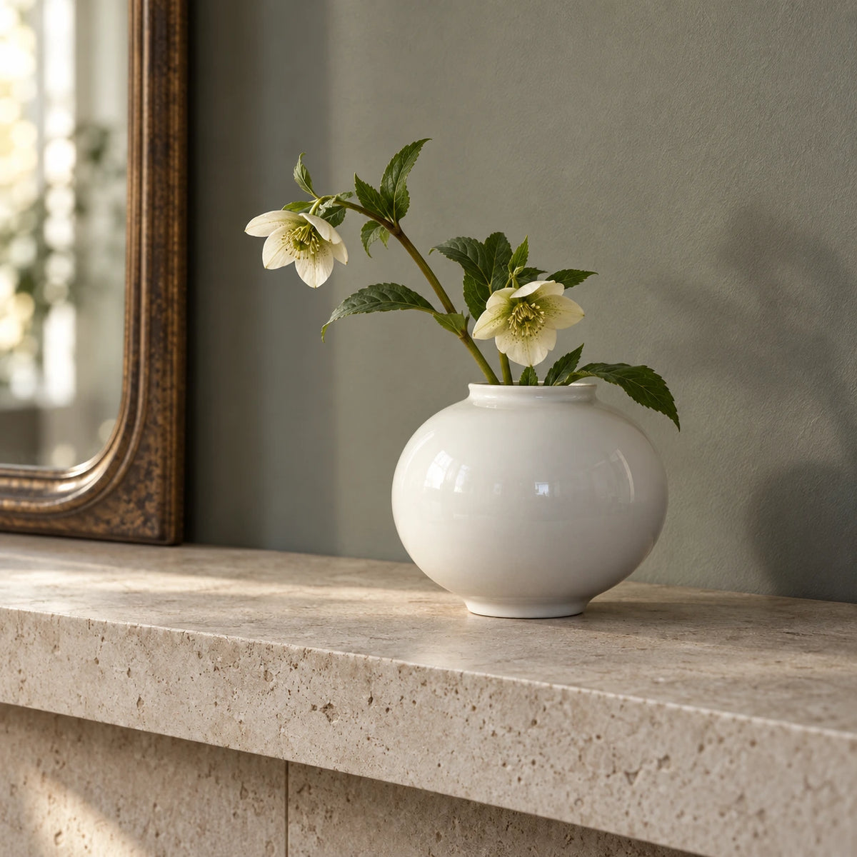 Small white moon vase with cream flowers and green leaves on a pale stone console beside a framed mirror.