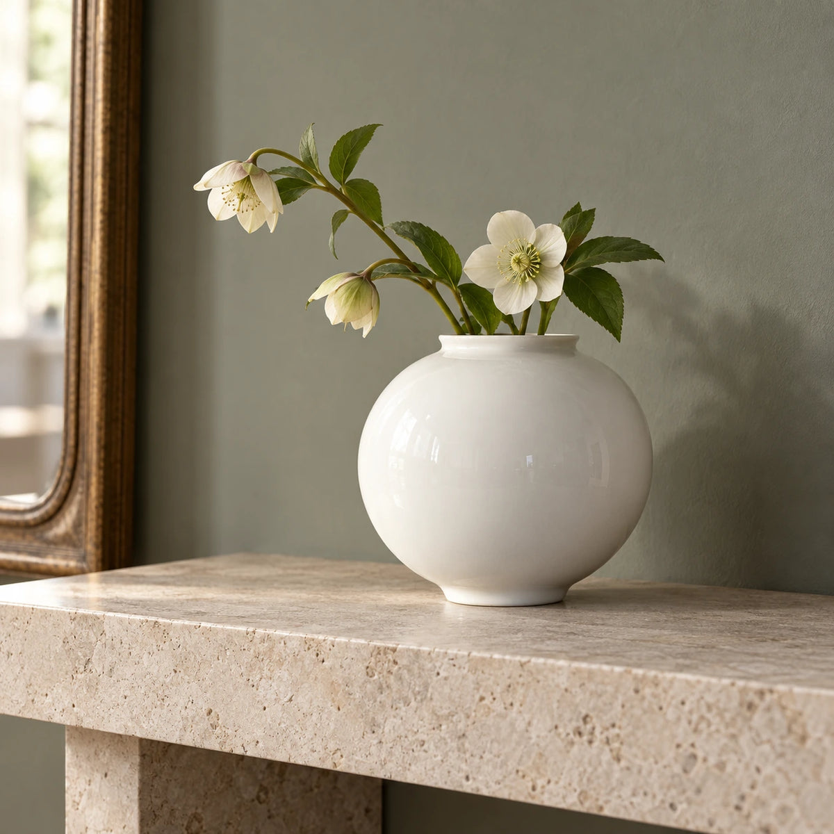 Medium white moon vase with cream flowers and green leaves on a pale stone console beside a framed mirror.