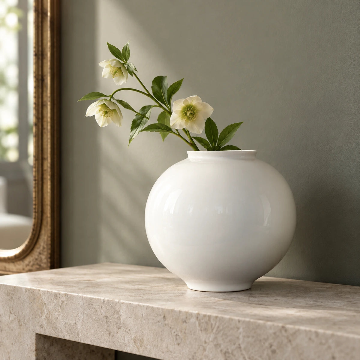 Large white moon vase with cream flowers and green leaves on a pale stone console beside a framed mirror.