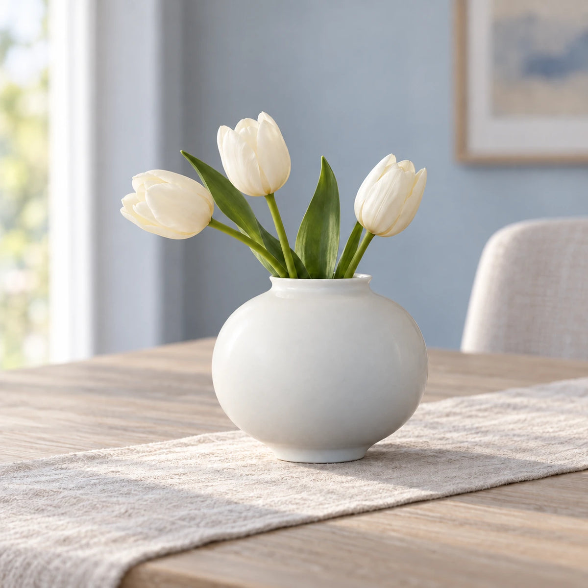 Kleine weiße Mondvase mit drei elfenbeinfarbenen Tulpen auf einem Leinenläufer auf einem hellen Esstisch.