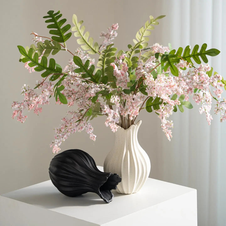 Ivory speckled fluted ceramic vase holding pink flowers, with a matching black ribbed vase on its side by a window.