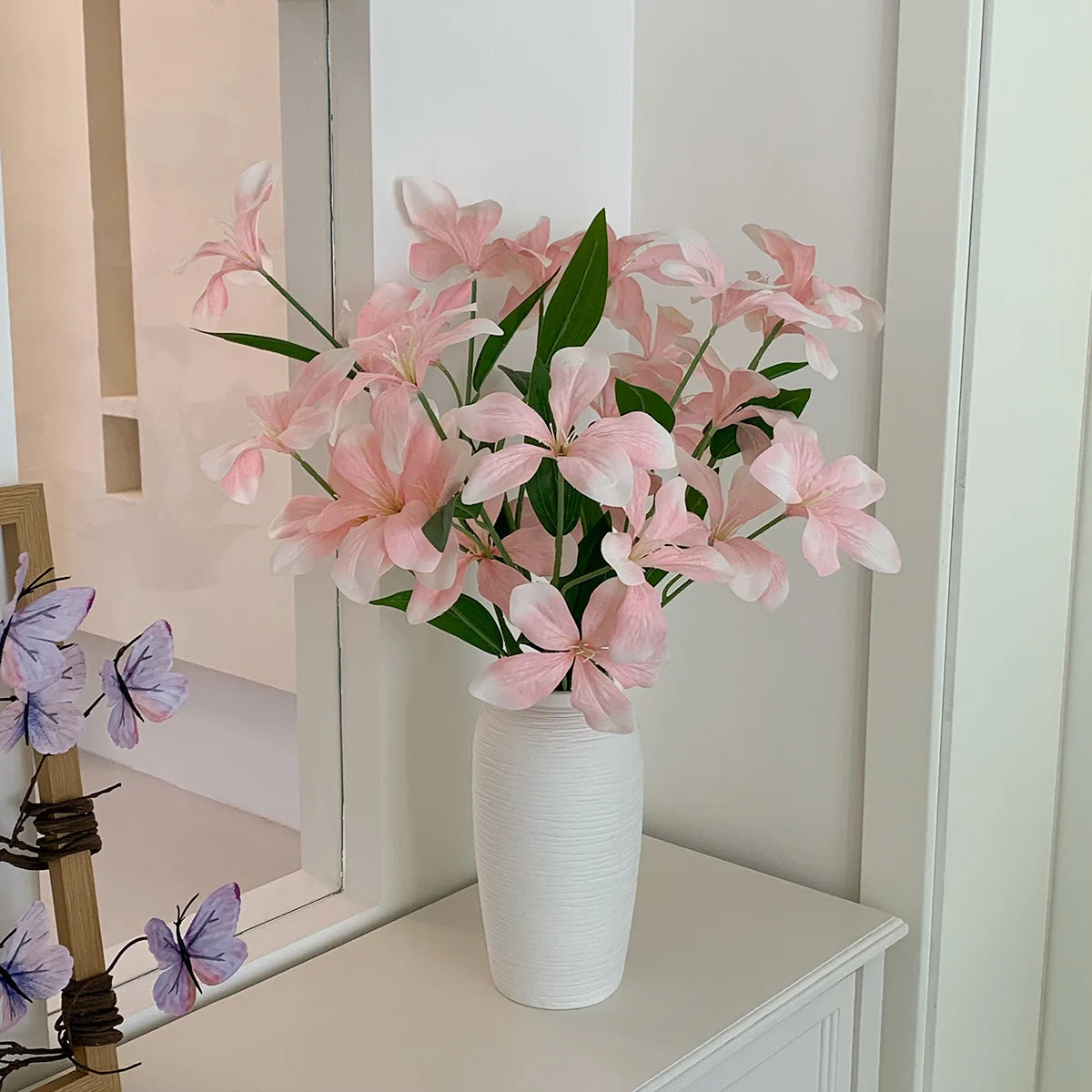 Pink artificial flowers with green leaves in a textured white vase on white table