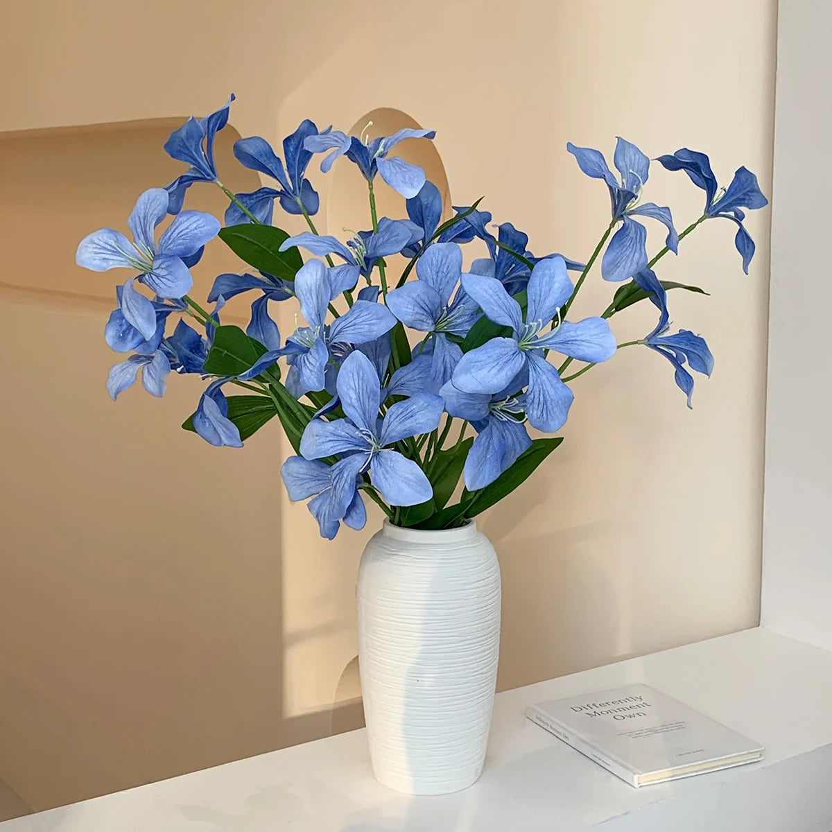 White ceramic vase with blue artificial flowers on white shelf near beige wall and book