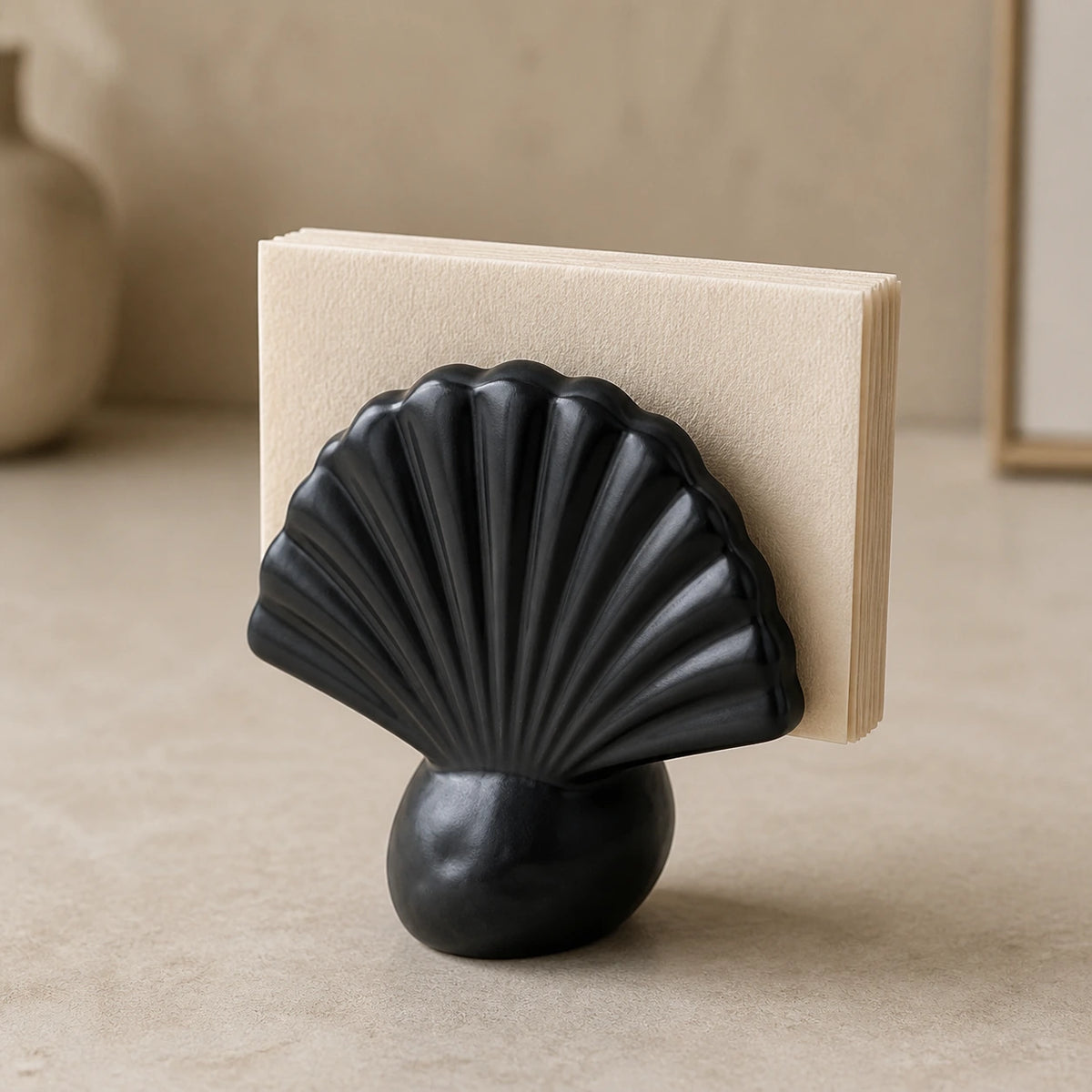 Schwarzer Keramik-Serviettenhalter in Muschelform mit gefalteten Papierservietten auf hellem Steintisch