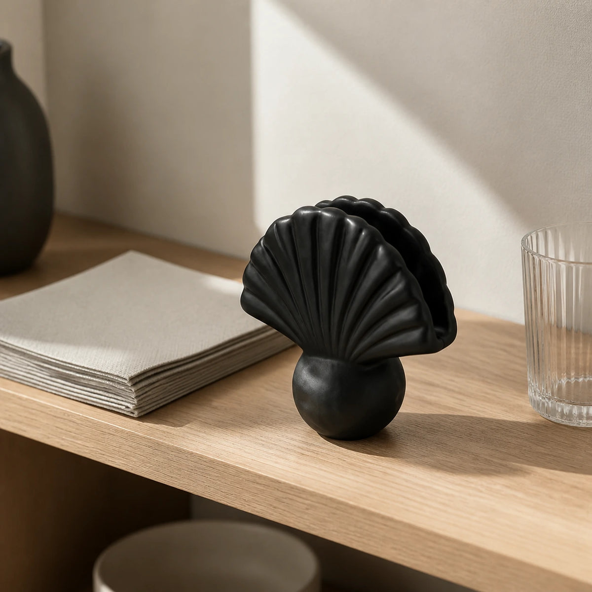 Schwarzer Keramik-Serviettenhalter in Muschelform neben gefalteten beigen Servietten und klarem Glas auf dunklem Holztisch