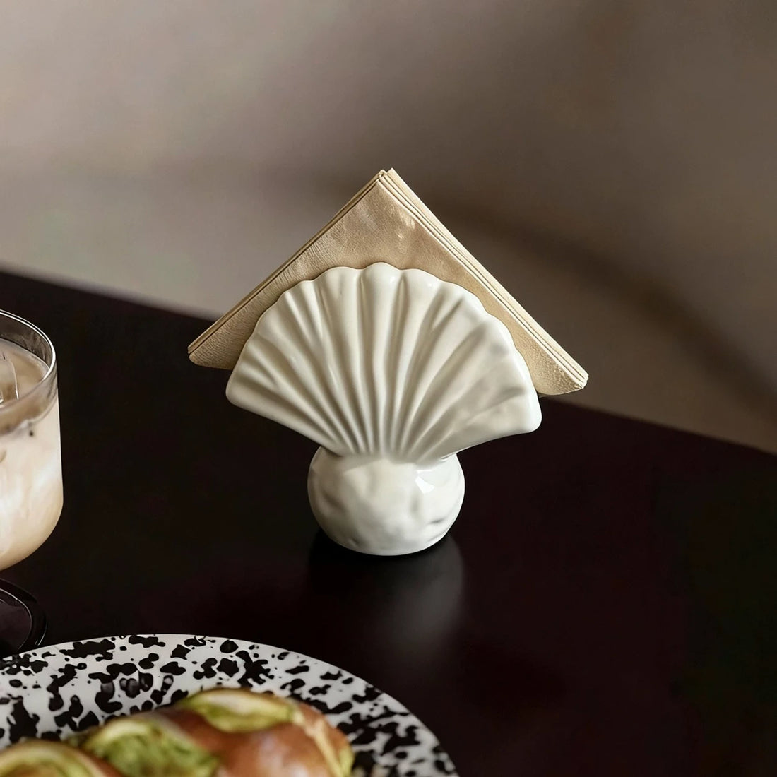 Warm ivory scallop shell ceramic napkin holder with folded beige napkins on a dark dining table