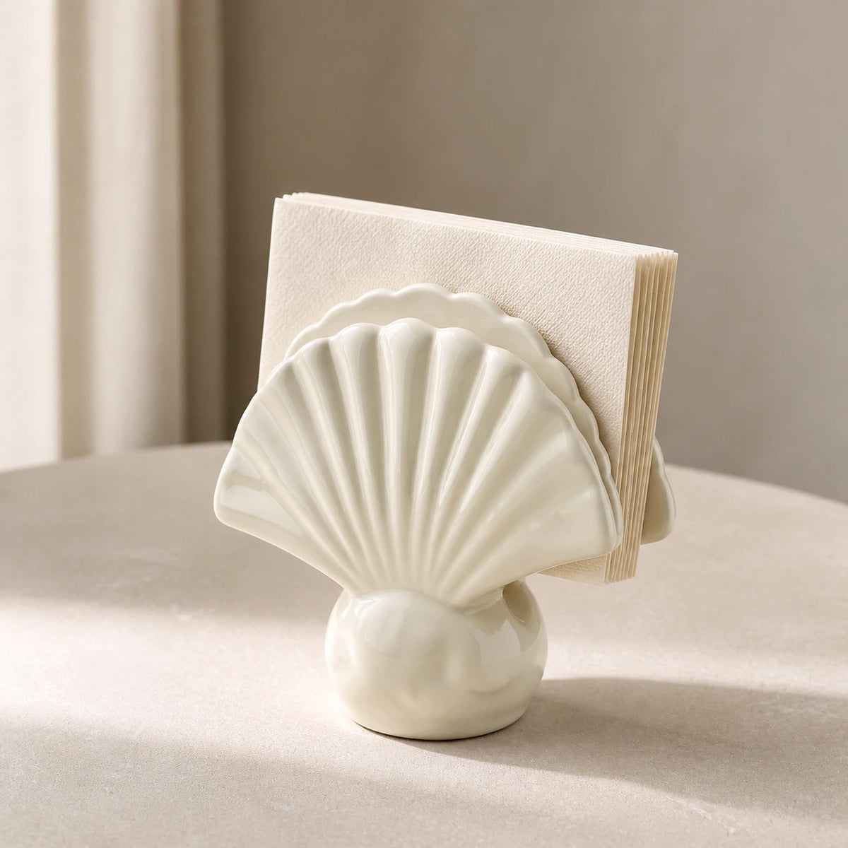 Elfenbeinfarbener Keramik-Serviettenhalter in Muschelform mit gefalteten Papierservietten auf hellem Steintisch