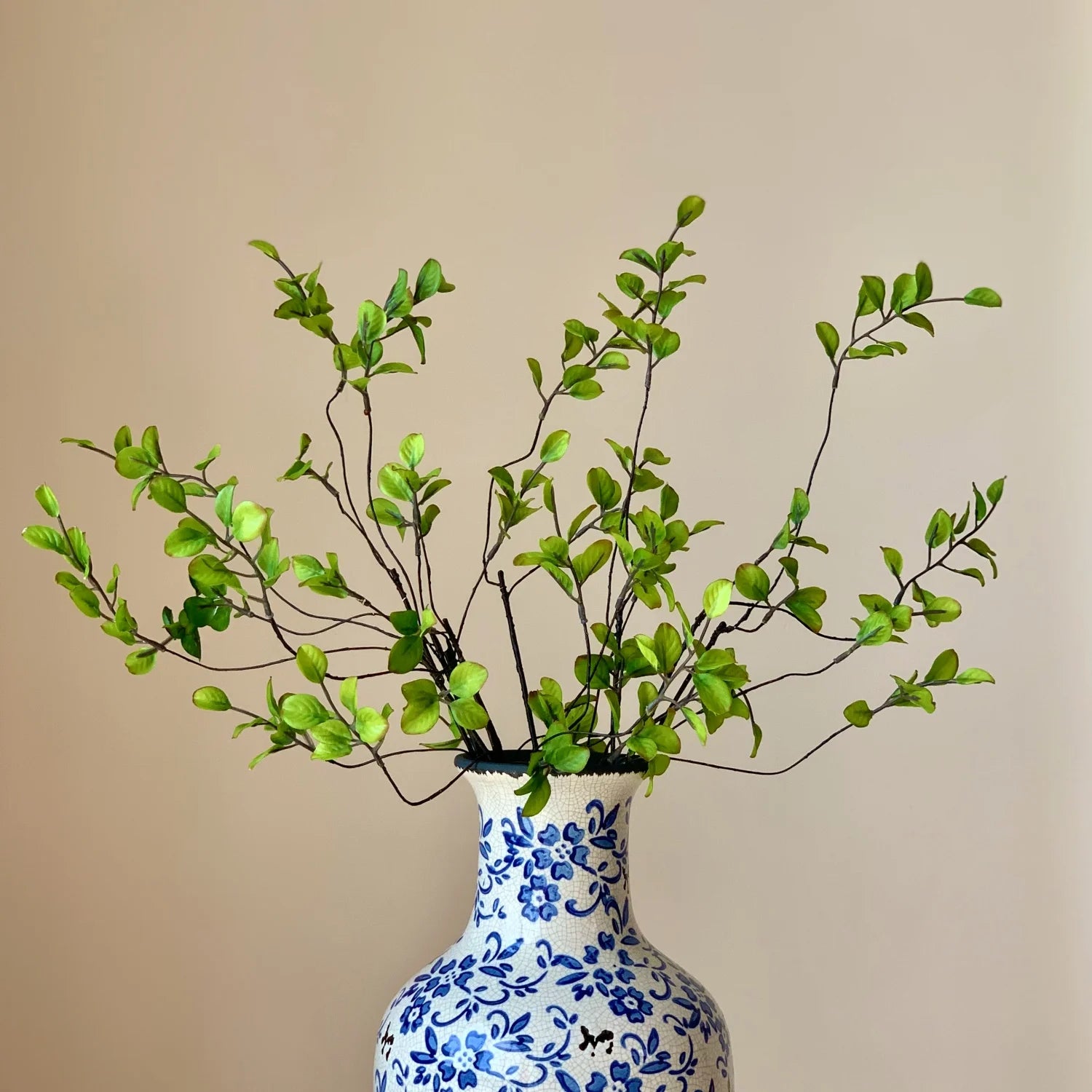 Blue and white floral ceramic vase with green leafy branches against beige wall