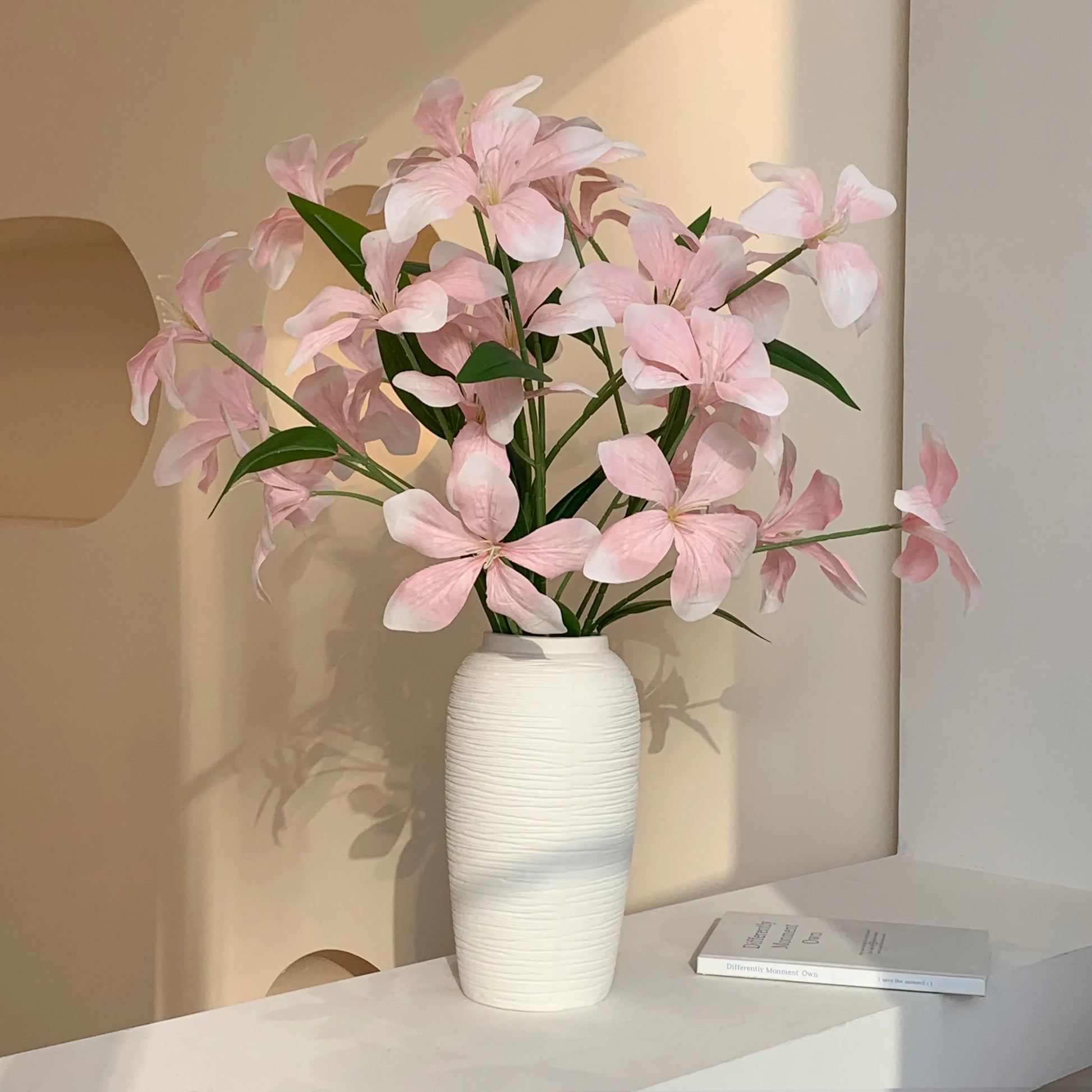 White ceramic vase with pink flowers and green leaves on a white shelf with book