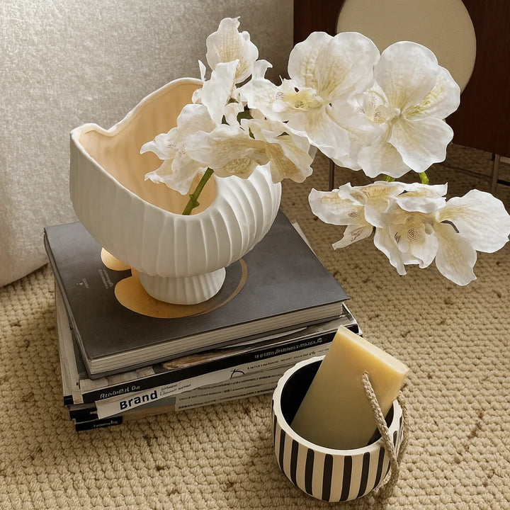 White fluted ceramic pedestal vase with an asymmetric wavy rim holding arching white orchids atop stacked books.