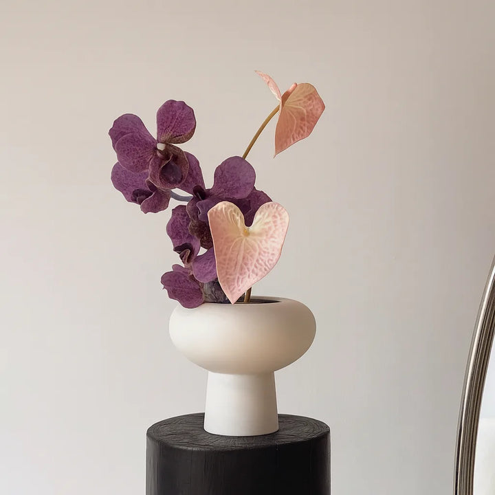 Faux floral bouquet with purple orchids and pink anthuriums in a white pedestal vase beside an arched mirror.