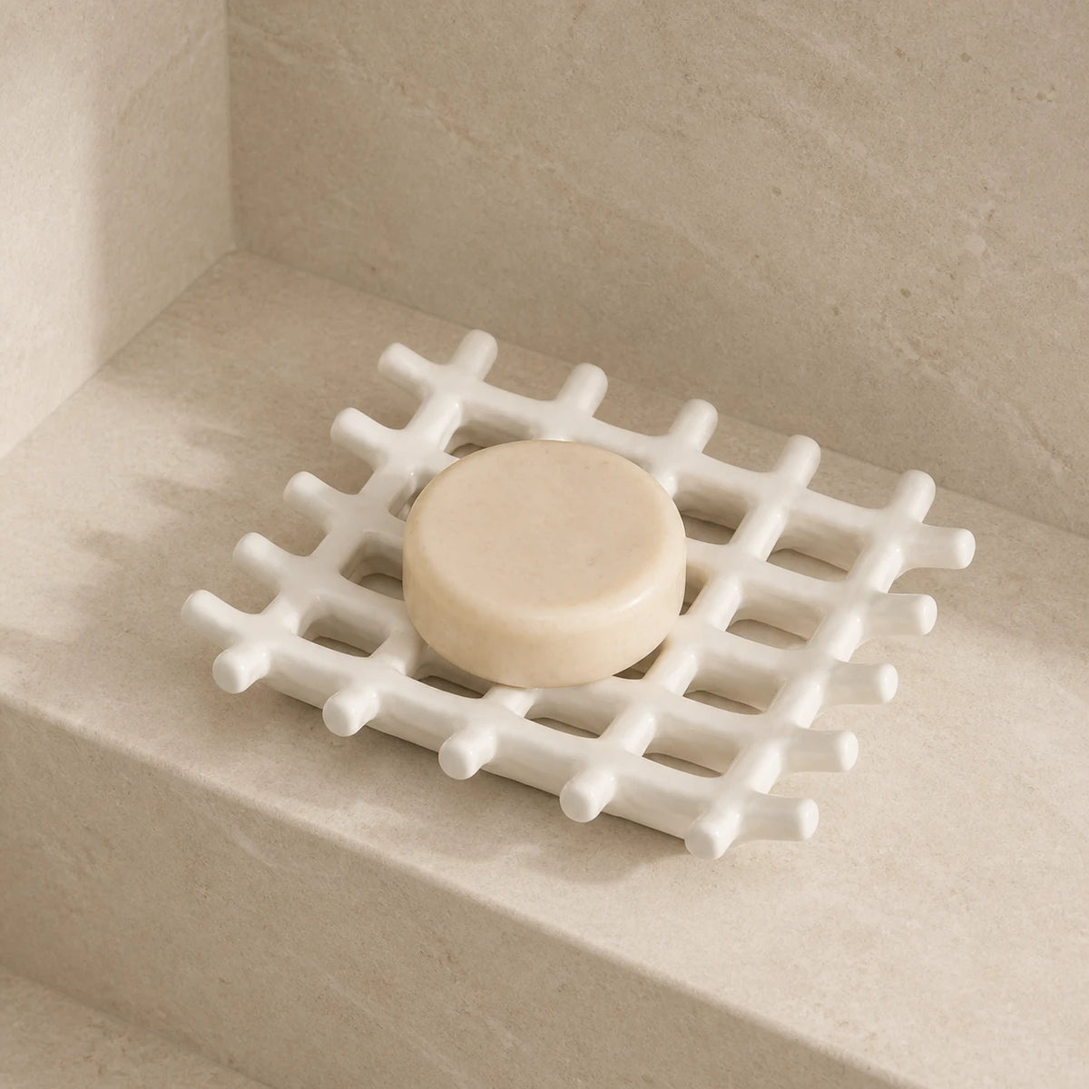 Porte-savon en treillis blanc mat avec savon rond sur un rebord en calcaire sous une douce lumière du soleil